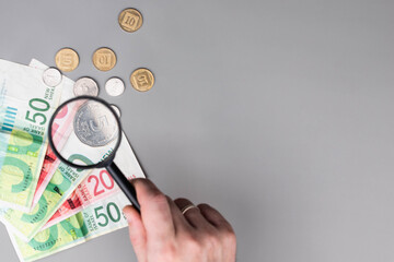 Israel currency 100 and 50 New Israeli shekel (NIS) banknotes and agorot coins under magnifying glass, grey background, concept of inflation, economic crisis, rising prices, and financial management.