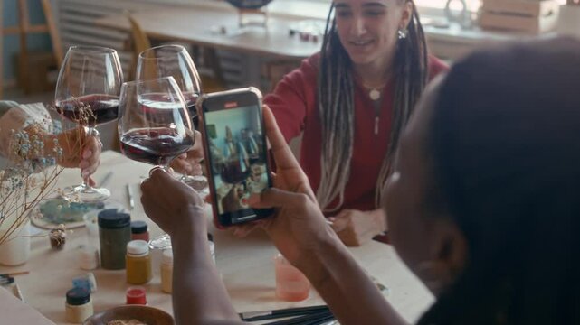 Slowmo shot of hands of girl holding smartphone recording vlog while clinking wine glasses with her girlfriends sitting at table with paint jars and brushes enjoying paint and sip workshop together