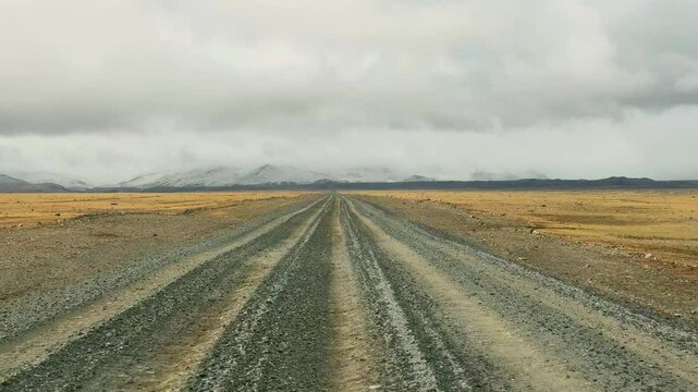 Gravel road stretching from Altai towards the Mongolian border with snowy mountain ridge. Desert steppe landscape under stormy cloudy sky showcasing wild remote terrain