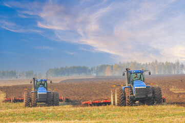 Obraz premium Two robust blue tractors cultivate a broad expanse of dark soil under swirling cloud formations, forging steady progress across open fields with autumn trees in the background.