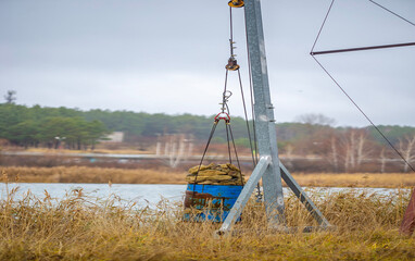 A makeshift crane hoists a blue metal barrel filled with stones near a marshy lakeshore. The simple contraption stands against a backdrop of reeds and distant pine trees.