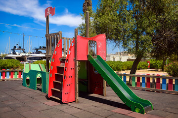 Small pirate-themed play structure with slide and climbing stairs in a sunny park. Surrounded by trees, colorful fence, and yachts in the background. Fun and safe place for children to play.