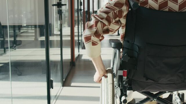 Back tracking midsection shot of anonymous office worker with disability wearing casual clothes, riding in wheelchair along long hallway, inclusivity and equal opportunities