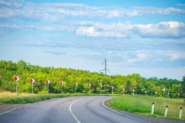 Bright red chevron signs line a sweeping curve in the road, surrounded by vivid green foliage under...
