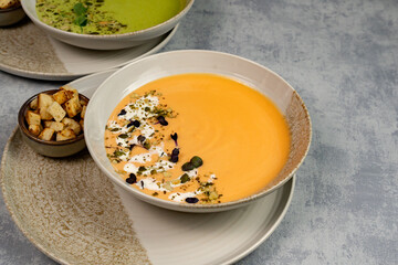 Bowl of pumpkin soup garnished with cream, pumpkin seeds and microgreens, with croutons next to it on a gray background, top view. Vegan or vegetarian food. Healthy dinner or lunch. Cooking.