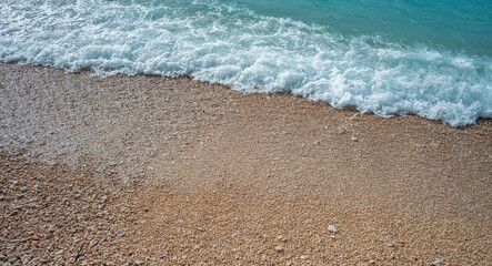 A gentle turquoise wave rolls onto a pebbled beach, creating white foam along the shoreline in a peaceful seaside scene.