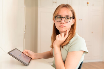 Teen girl with glasses holds a tablet and rests her chin on her hand, sitting at a desk in a bright...