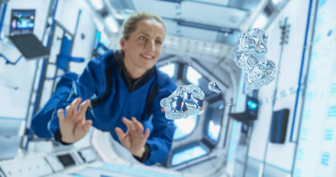 Female Cosmonaut in a Blue Suit Interacts With Floating Water Droplets Inside a Space Station, Demonstrating Gravity Effects. Portrait of a Courageous Female Astronaut on Board a Spacecraft, Floating