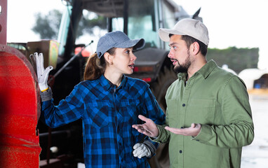 Man and woman farmers standing at tractor in garage space and talking.