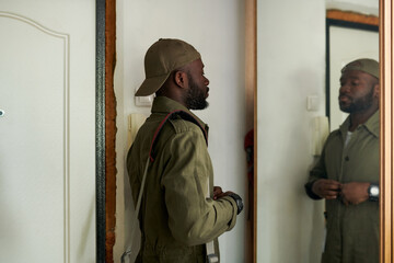 Black man in green uniform standing in front of mirror, getting ready for work. Wall decor and cap visible, creating a thoughtful and introspective scene