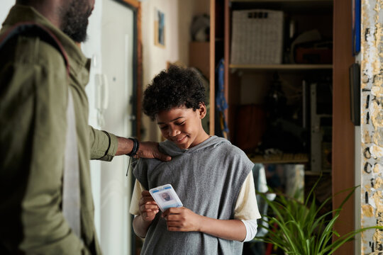 Teen boy smiling while holding identification card close to chest, engaging with unseen dad. Setting includes home interior with plants and furniture visible
