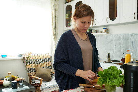 Woman preparing vegetables while cooking meal in a cozy kitchen with various utensils and fresh produce around. Wearing comfortable clothes, engaging in healthy lifestyle