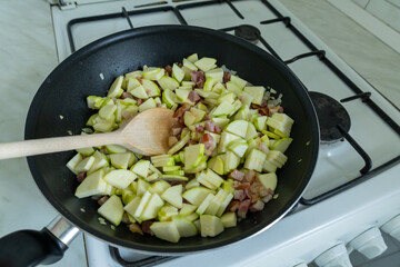 Overhead view of chopped zucchini and bacon being sautéed in a black frying pan on a gas stove. 