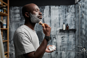Black person shaving face with shaving foam in modern, stylish bathroom with a textured wall background showing personal grooming routine and attention to detail