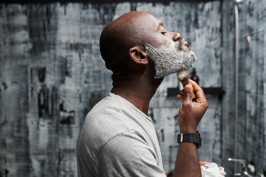 Black man with beard shaving in front of mirror in modern bathroom, using shaving cream and razor. Man is taking care of grooming and hygiene without looking at camera