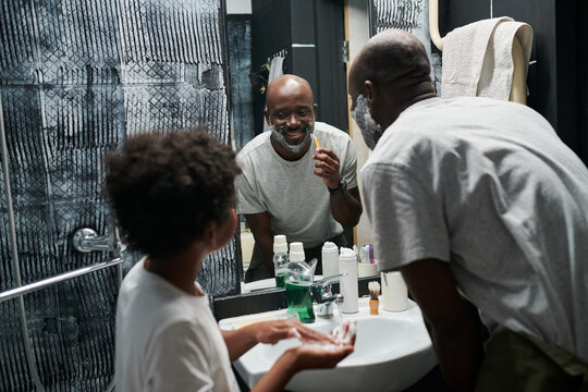 African American man shaving his face while talking with child in modern bathroom with stylish decor. Father and child bonding moment