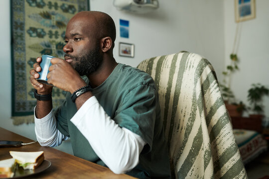Man savoring hot drink while seated at wooden table in warmly decorated room with plants and wall hangings around him