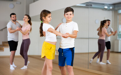 Obraz premium Young girl and boy dancing waltz together during group family training. Their parents dancing in background.
