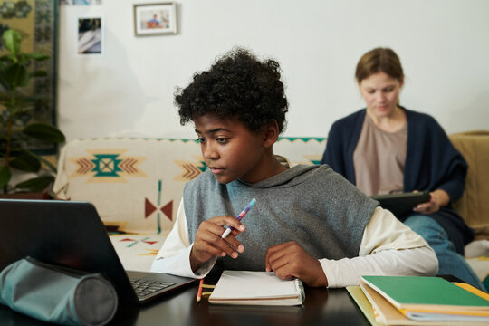 Young boy sitting at a table with laptop and notebook, concentrating on studies while holding a pen. Woman in background using tablet, creating cozy atmosphere - Powered by Adobe