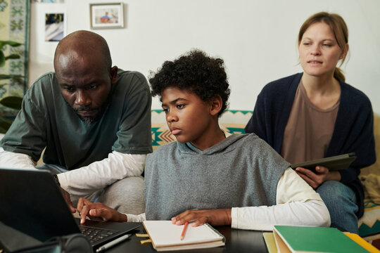 Family consisting of African American father, biracial child, and Caucasian mother working on childs homework using laptop and notebook while sitting on couch