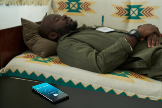 Man lying on bed with phone showing alarm on screen foregrounded on table. Setting seems casual and relaxed, suggesting a peaceful indoor environment