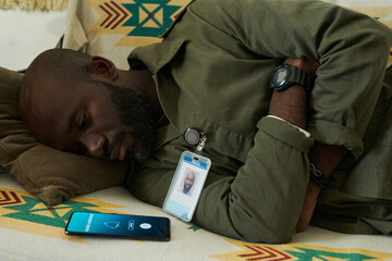 Man sleeping on couch with head resting on cushion, arm bent under his head, and an ID badge hanging from his shirt, smartphone placed beside him