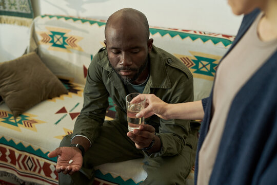 African American man in casual clothing seated on sofa receiving medication and water from another person