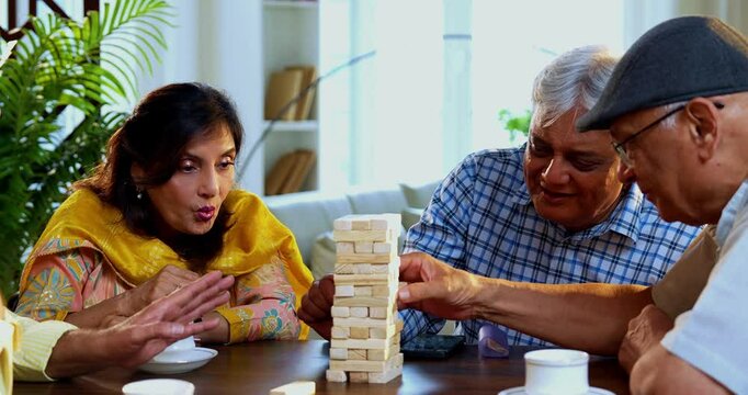 Indian senior adults playing jenga game or wooden block balancing game while seated around dining table in cozy Indian home, laughing, celebrating fun moments, bonding, enjoying friendly reunion time