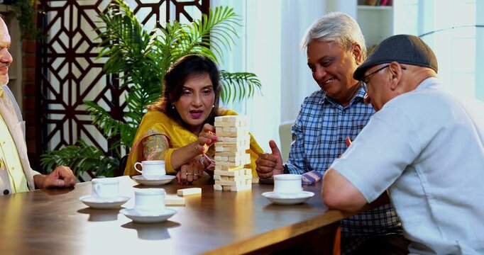 Indian senior adults playing jenga game or wooden block balancing game while seated around dining table in cozy Indian home, laughing, celebrating fun moments, bonding, enjoying friendly reunion time