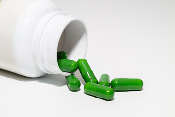 Green capsules spill from a white medicine bottle onto a white surface, suggesting health or herbal supplements.
