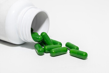 Green capsules spill from a white medicine bottle onto a white surface, suggesting health or herbal supplements.