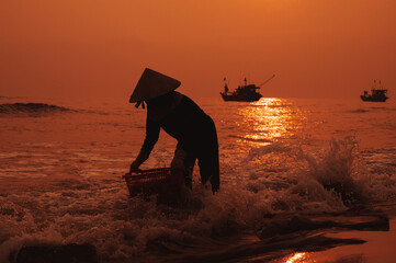 Silhouette of a Vietnamese woman in a straw hat on the shore of sea with ships in a fishing village in the morning at sunrise in Vietnam