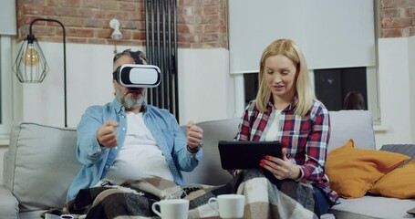 Man in special 3d glasses as if behind the wheel while his smiling cute wife sitting near him on comfortable sofa and choosing game for him on tablet pc - Powered by Adobe