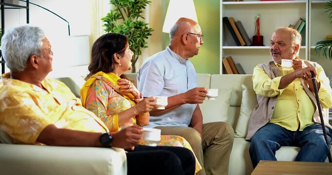 Seniors drinking hot tea coffee while seated on cozy sofa as Indian Asian old age friends enjoy warm beverages together at home during cheerful reunion moment in a relaxed modern indoor setup