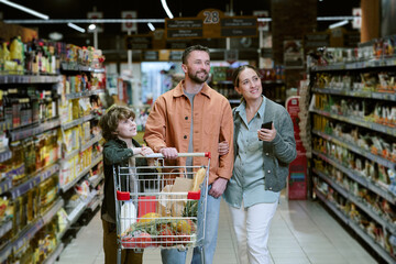 Family enjoying grocery shopping in supermarket, with young child pushing cart filled with various groceries while parents look around and smile, capturing fun bonding moment © AnnaStills