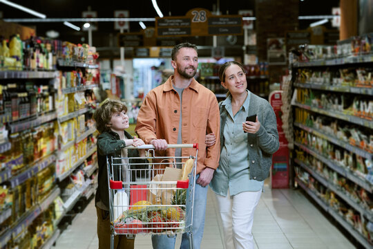 Family enjoying grocery shopping in supermarket, with young child pushing cart filled with various groceries while parents look around and smile, capturing fun bonding moment
