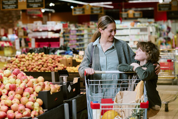 Smiling woman pushing shopping cart with happy child embracing her in grocery store produce section...
