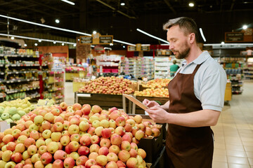Caucasian male worker in apron examining fresh apples in grocery store produce section while holding tablet. Display of different fresh fruits in background
