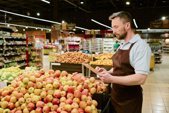 Caucasian male worker in apron examining fresh apples in grocery store produce section while holding tablet. Display of different fresh fruits in background