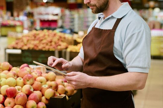 Grocery store worker wearing brown apron holding tablet near apples, demonstrating usage of digital technology in a vibrant supermarket setting filled with fresh produce - Powered by Adobe