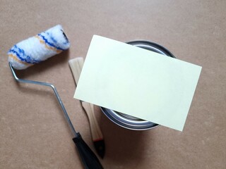 Blank sheet of paper on closed tin lid of paint can. Closed paint can and roller with brush on brown background