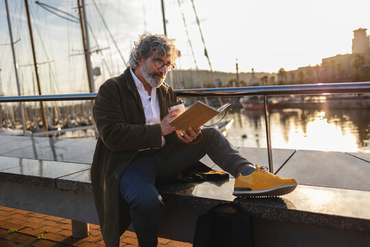 Professor enjoying book and coffee by city harbor at sunset - Powered by Adobe