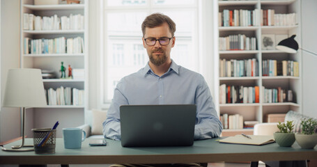 Creative Man Works on His Laptop at a Desk. Bright Room, Filled With Bookshelves. Professional...