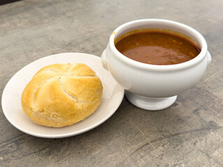 Rustic soup in white bowl with crusty bread roll on plate