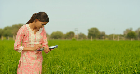Indian young adult farmer woman lady wear casual dress desi life outdoor holding tablet device website looking screen asian smiling teen age girl use tab yield data outside work rural area day time