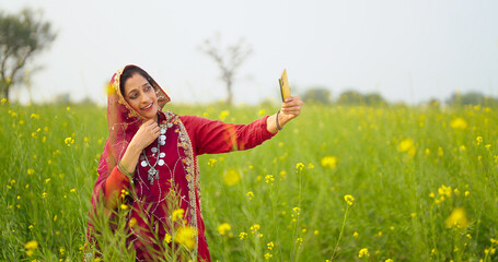 Indian adult gen x farmer woman lady wear red saree standing outdoor field hold mobile take selfie...