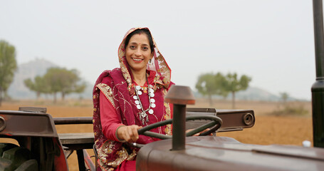 Indian adult woman lady smiling sitting on tractor transportation look at camera outdoor. Asian alone village farmer mother wear red traditional saree do pose staring cam outside enjoy day © Raushan_films