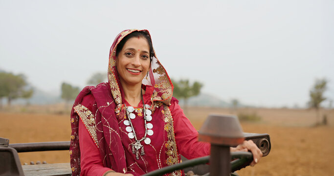 Indian adult woman lady smiling sitting on tractor transportation look at camera outdoor. Asian alone village farmer mother wear red traditional saree do pose staring cam outside enjoy day