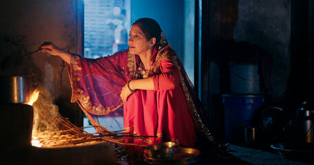 Indian villager gen x woman preparing food using traditional earthen mud stove indoor house asian young female wear red saree cooking on open fire flam inside kitchen home