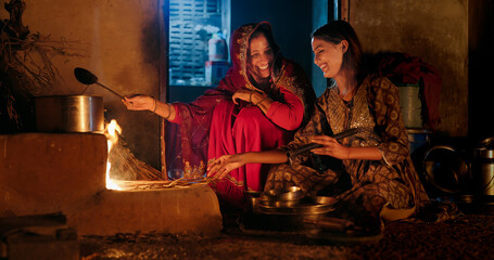 Indian villager mom with teen age female girl prepare food using earthen clay mud stove indoor house asian young adult woman lady wear red saree cooking on open fire flam inside kitchen home desi life